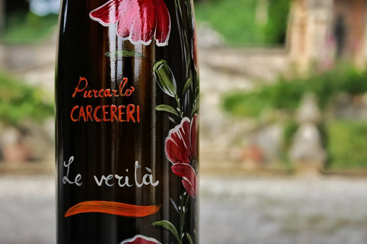 Close-up of a dark wine bottle with 'Piercarlo CARCERERI' and 'Le verità' hand-painted with red poppy flowers.