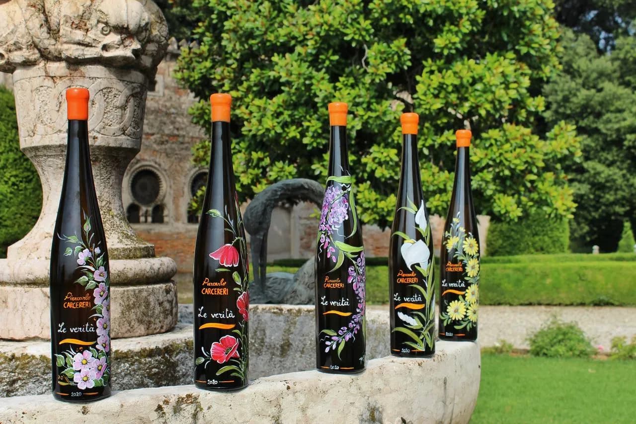 Five hand-painted wine bottles from the 'Piercarlo CARCERERI Le verità' series, featuring various floral designs, set on a stone ledge in a garden.