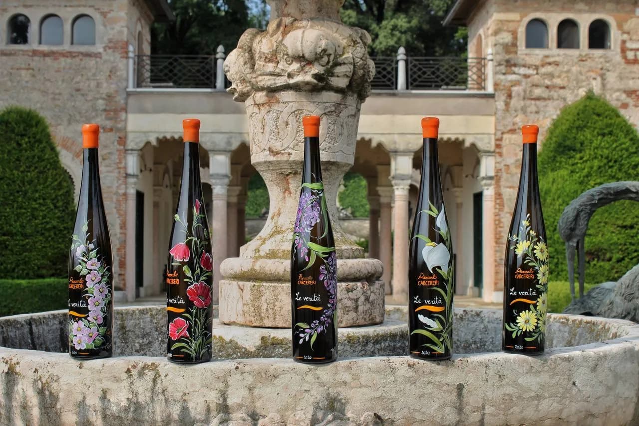 Five hand-painted wine bottles from the 'Piercarlo CARCERERI Le verità' series, each with a different floral design, set around a stone fountain.