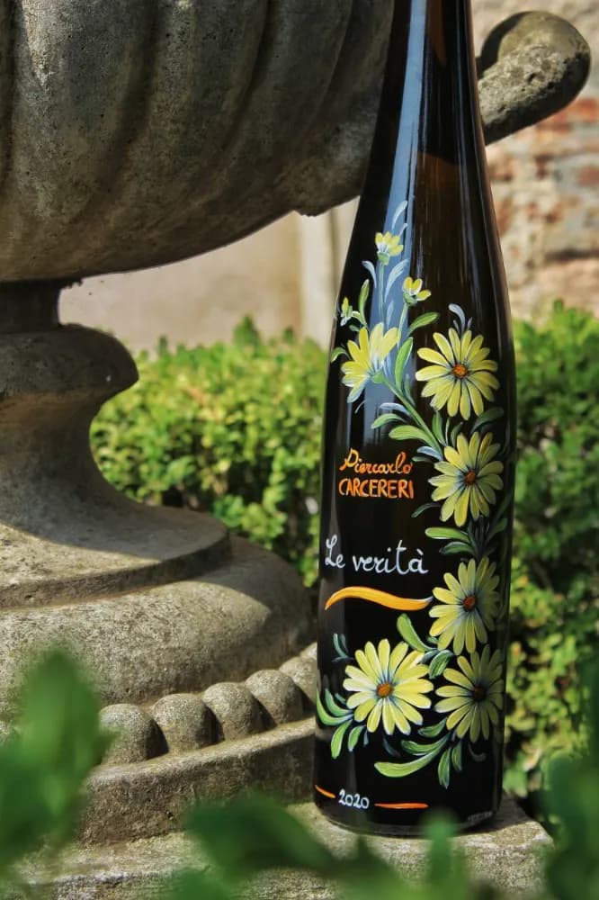Close-up of a dark wine bottle hand-painted with yellow daisies, labeled 'Piercarlo CARCERERI Le verità 2020', next to a stone fountain.