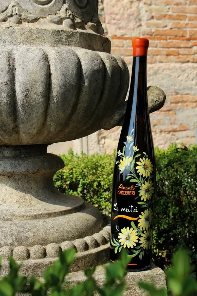A dark wine bottle hand-painted with yellow daisy flowers, labeled 'Piercarlo CARCERERI Le verità', resting against a weathered stone fountain in a garden.