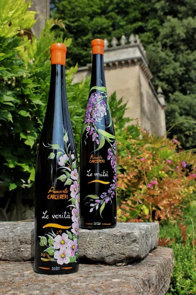 Two dark wine bottles, one with pink blossoms and one with purple wisteria, from the 'Piercarlo CARCERERI Le verità 2020' series, placed on a stone wall outdoors.